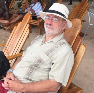 An older man with a beard and glasses wearing a hat and a light-colored shirt sitting in a wooden chair on a sandy beach.